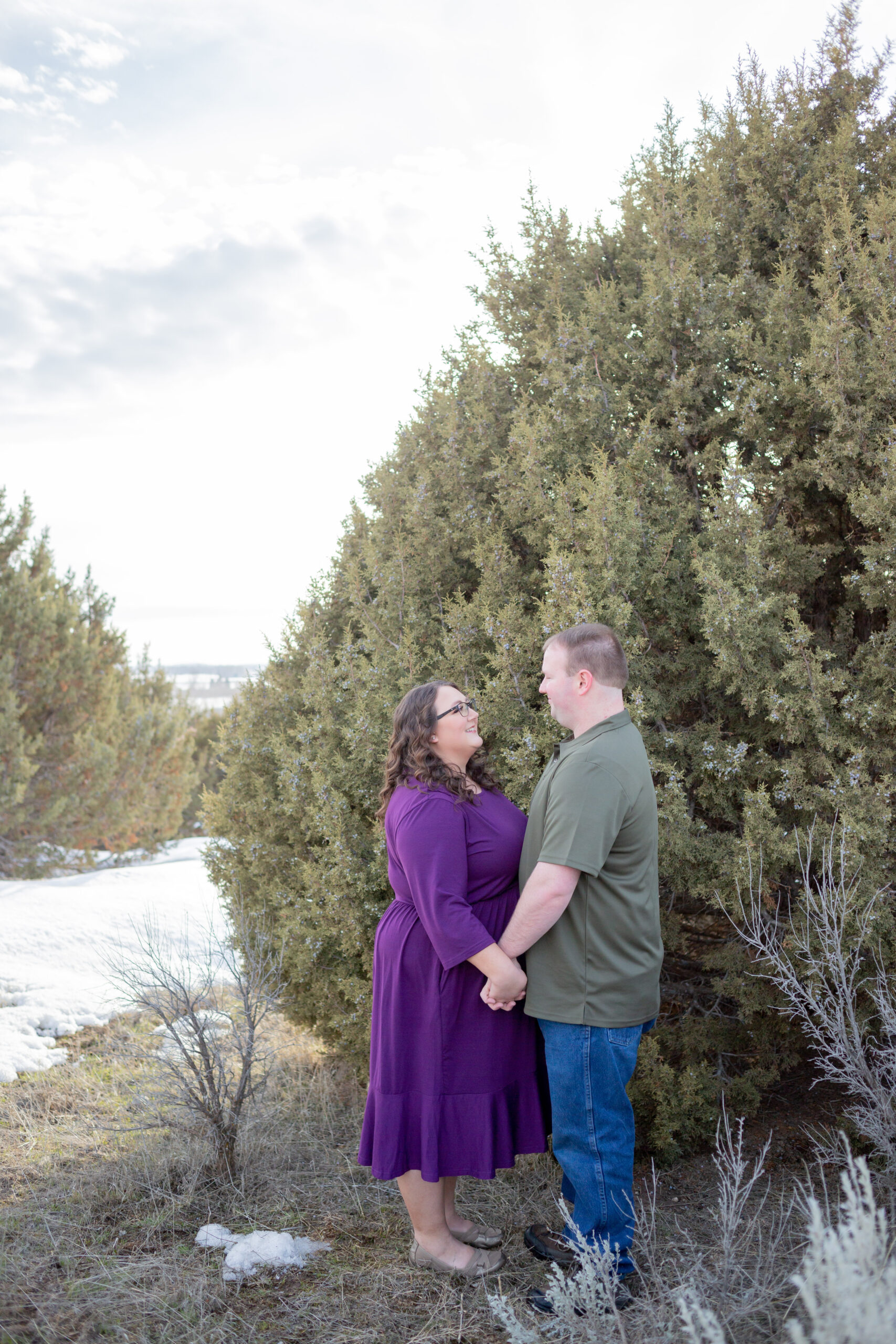 engaged-couple-standing-looking-at-each-other-with-cedar-trees-in-the-background