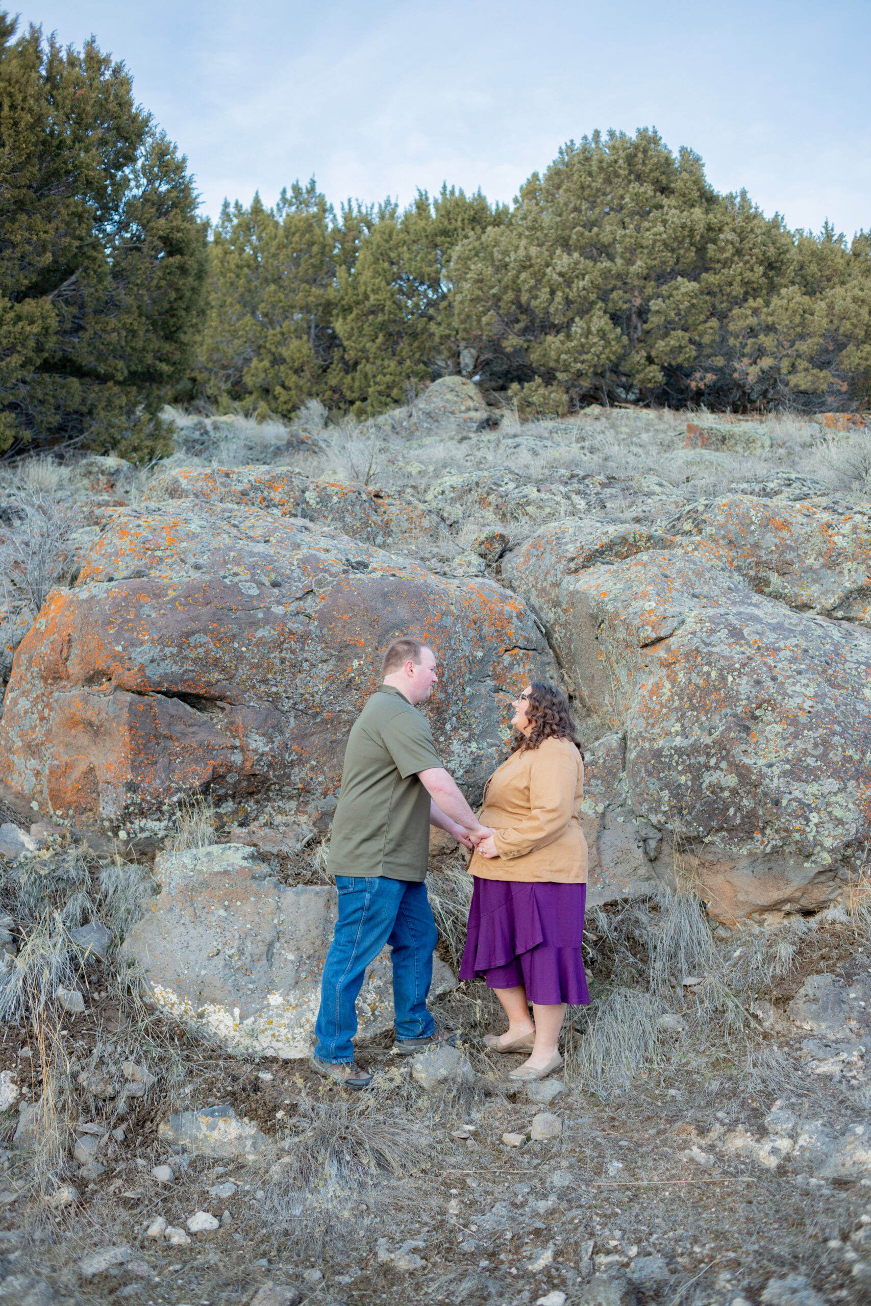engaged-couple-standing-looking-at-each-other-with-rock-cliff-and-trees-in-the-background